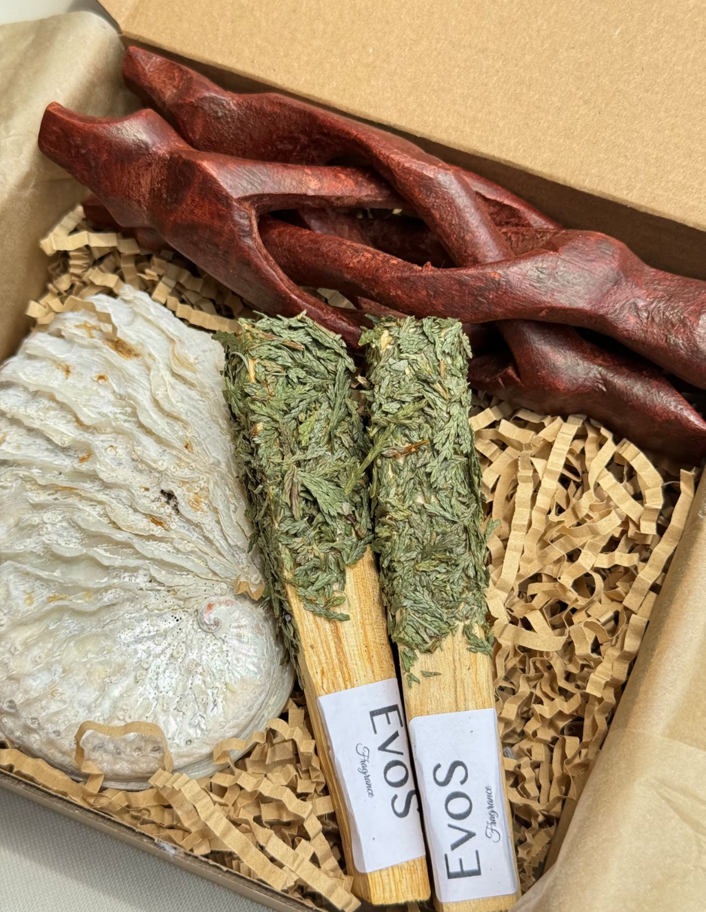 Vue rapprochée du coffret EVOS Fragrance avec deux bâtons de palo santo aux herbes séchées vertes, racine en bois rouge sculpté et savon nacré blanc, présentation sur papier kraft crêpé
