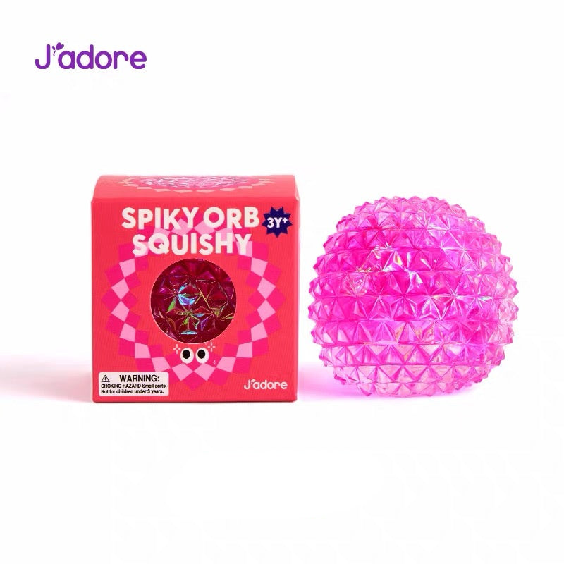 Balle squishy anti-stress J'adore Spiky Orb rose fuchsia irisé à texture picots avec sa boîte d'emballage damier, photo produit sur fond rose
