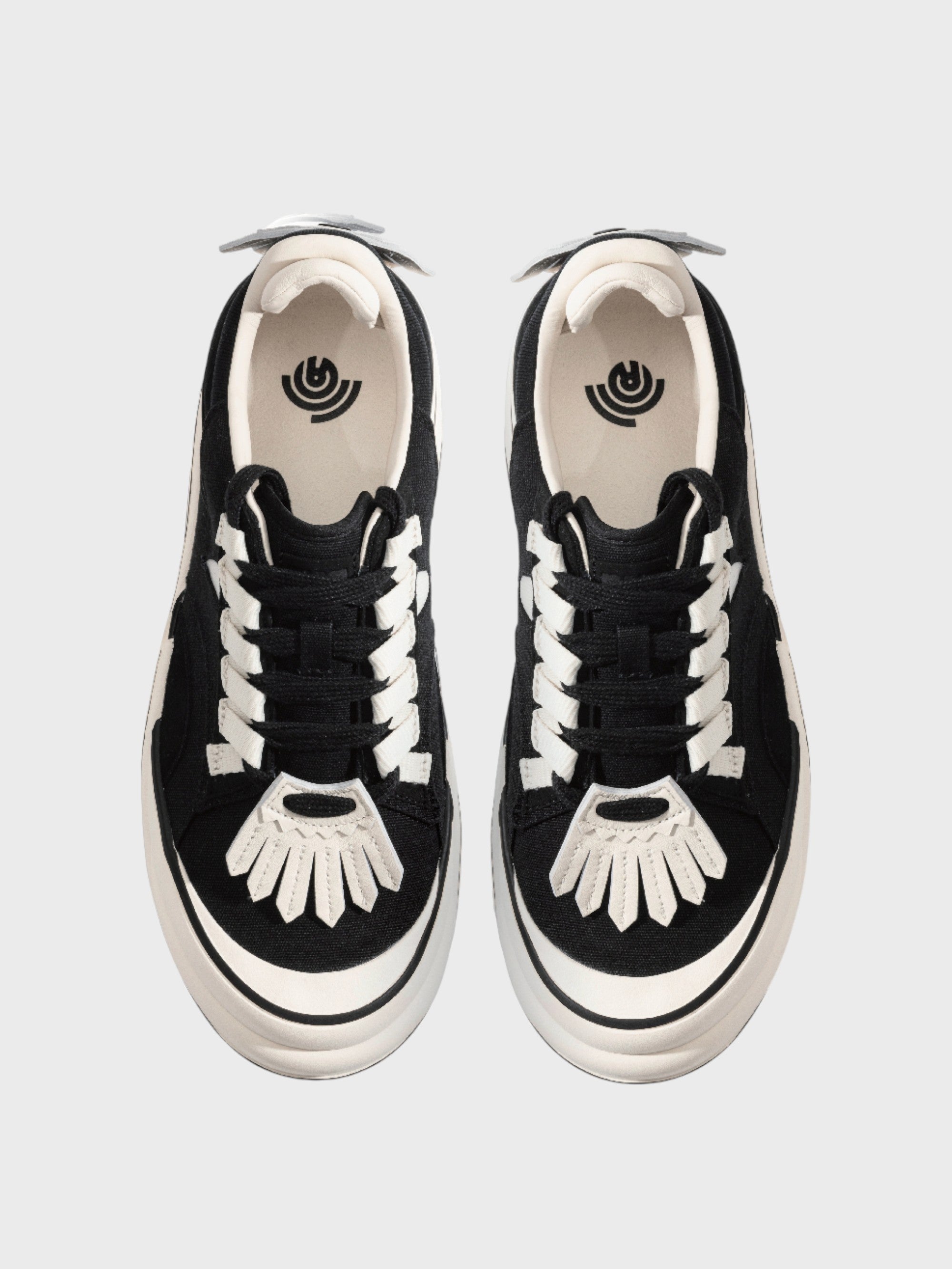 Radar Homing Beacon RHB sneaker – paire de sneakers daim noir lacets blancs plats, patch bout de pied blanc motif soleil éventail, logo œil concentrique intérieur, languette blanche, vue de dessus, fond gris clair
