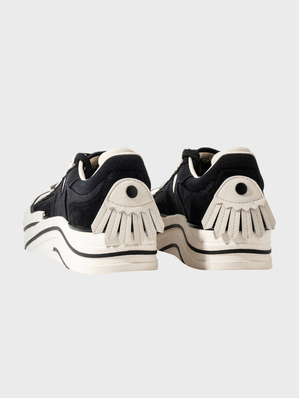 Radar Homing Beacon RHB sneaker – paire de sneakers daim noir semelle épaisse blanche, patch talon blanc motif œil soleil éventail avec point central noir coutures apparentes, vue de derrière, fond gris clair
