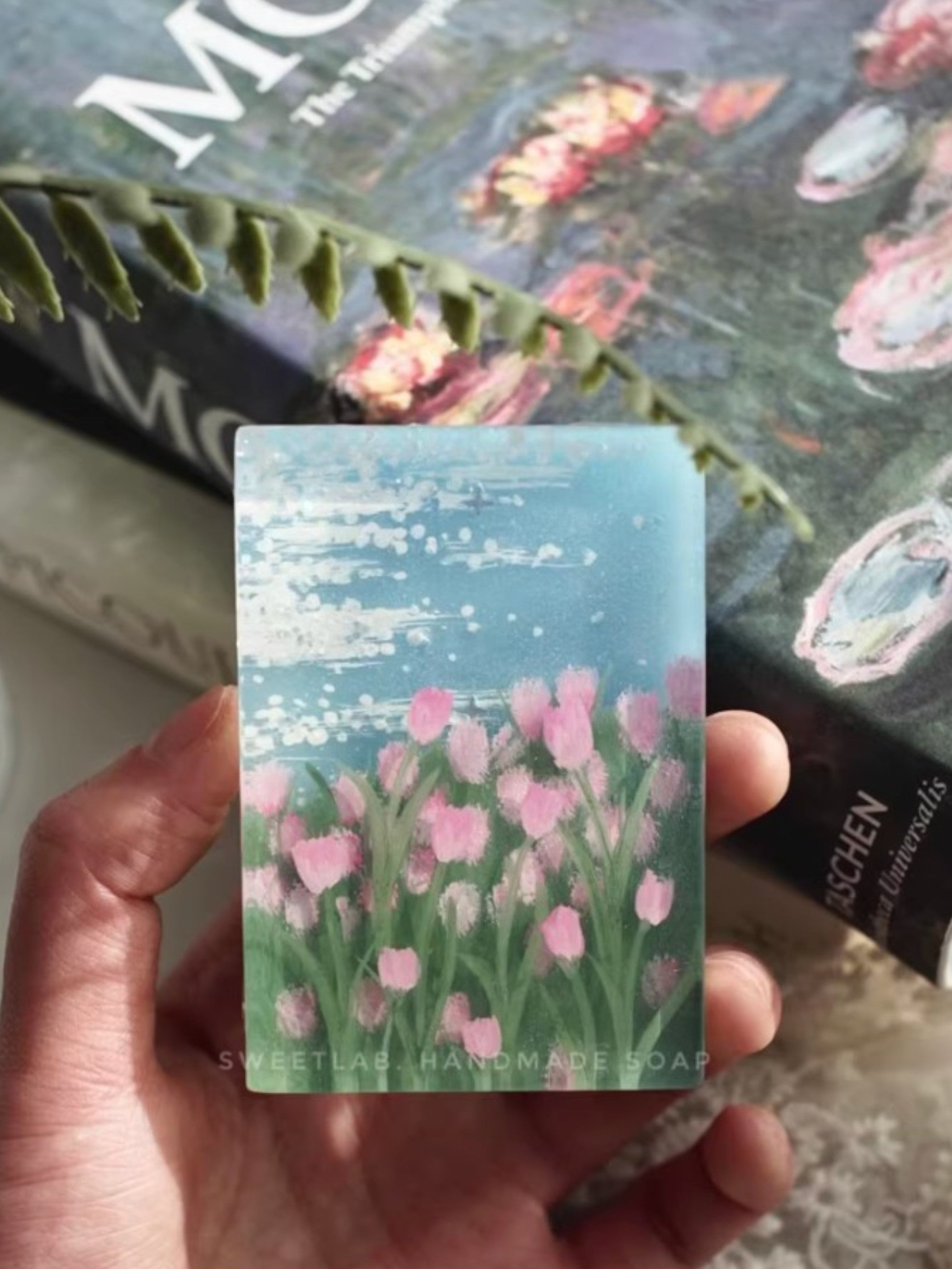 Savon artisanal Sweetlab Handmade Soap motif peinture champ de tulipes roses au bord d'un lac, tenu en main, photo lifestyle avec livres en arrière-plan
