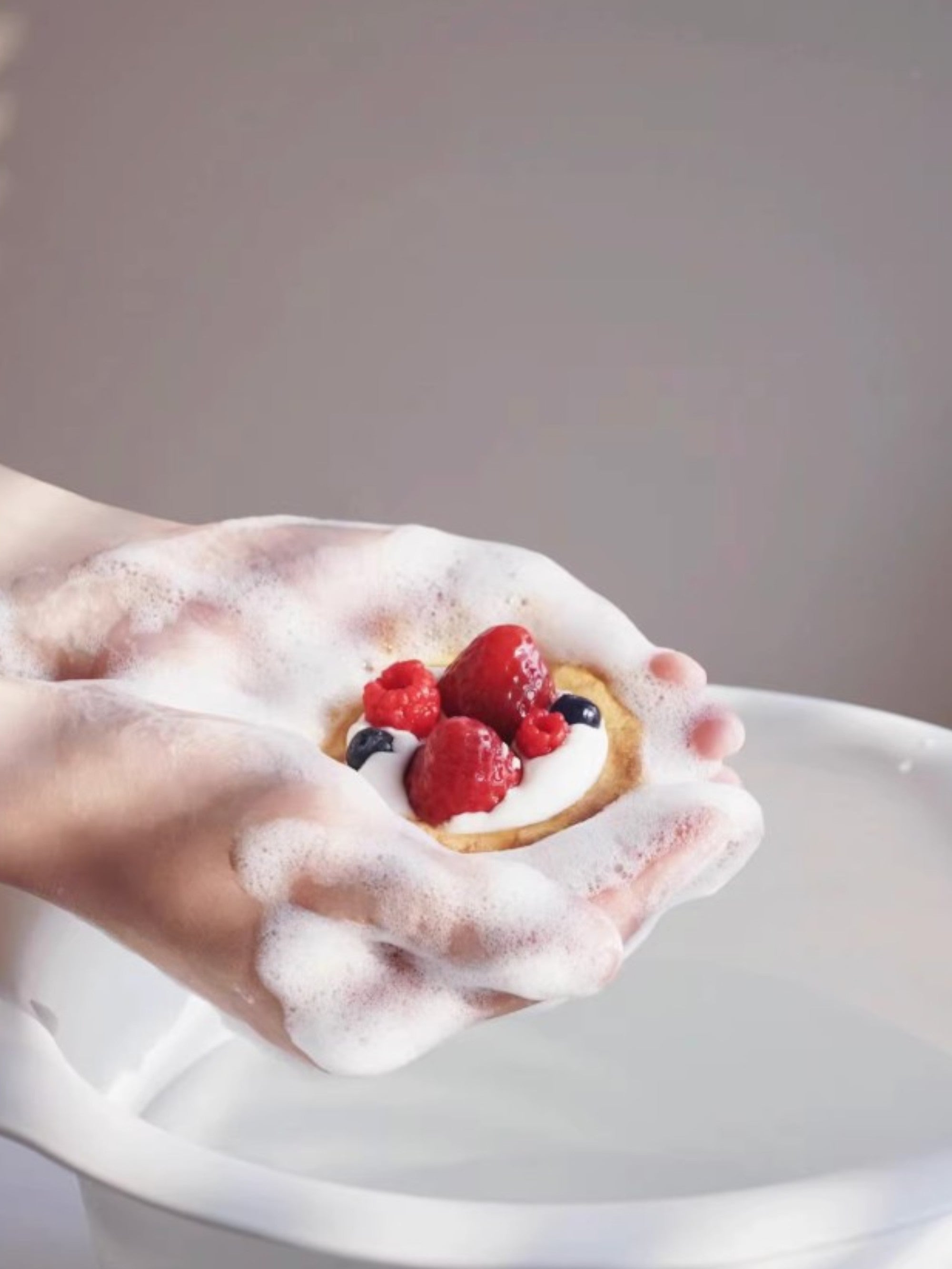 Savon artisanal Sweetlab en forme de tarte aux fraises, framboises et myrtilles, démonstration de lavage avec mousse dans la main, fond gris
