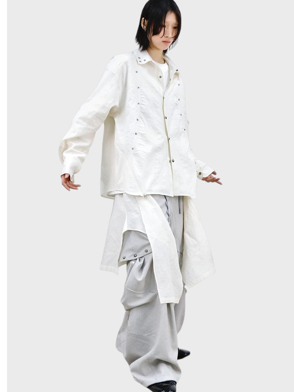 Chemise oversize RELABEL blanc cassé rivets portée par une femme, look complet avec pantalon ballon gris déconstructiviste et derbies noirs
