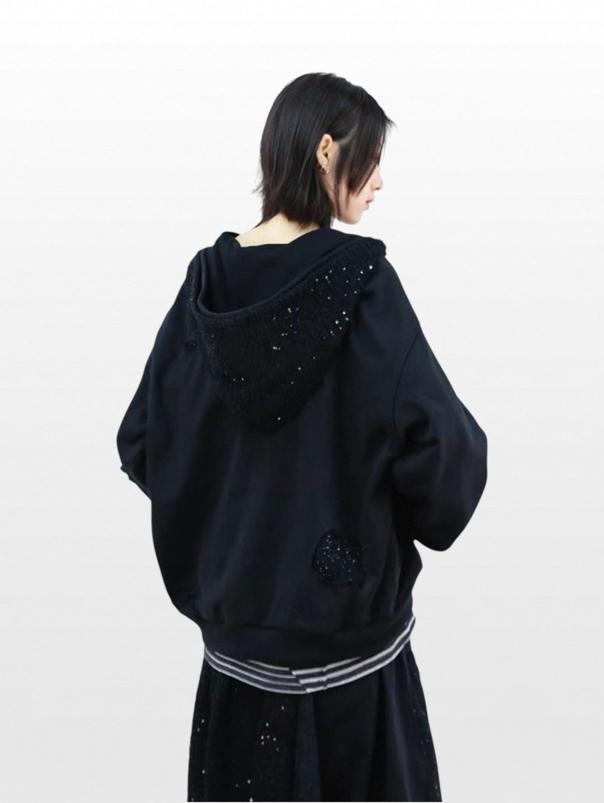 Hoodie zip oversize RELABEL noir strass porté par une femme, vue de dos avec incrustations strass capuche et bord-côte rayé gris, jupe large noire
