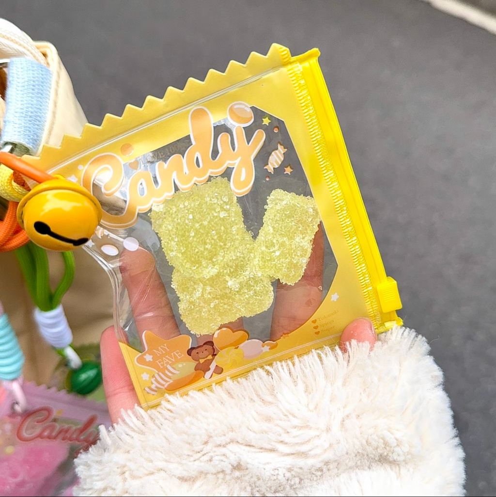 Jouet anti-stress ourson ananas sable scintillant jaune-vert tenu dans une main gantée blanc moelleux, sachet jaune vif Candy zip jaune avec grelot doré et cordon orange, ambiance kawaii rue
