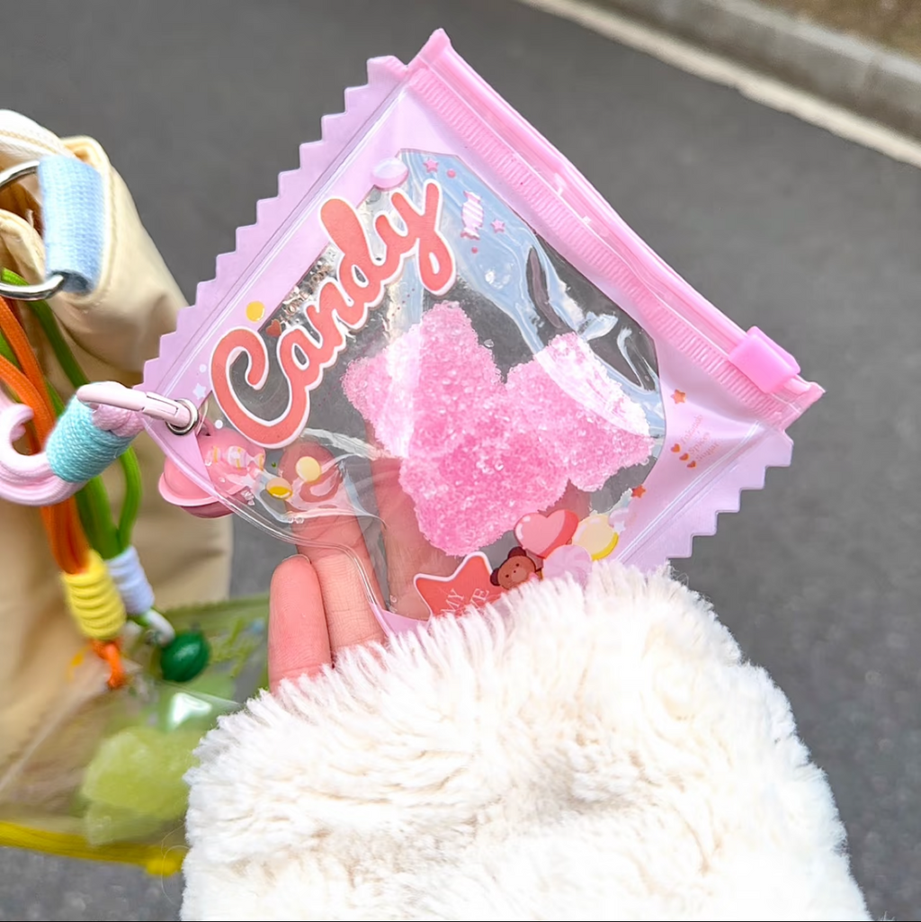 Jouet anti-stress ourson fraise sable scintillant rose tenu dans une main gantée blanc moelleux, sachet iridescent rose Candy zip rose avec confettis colorés, ambiance kawaii rue