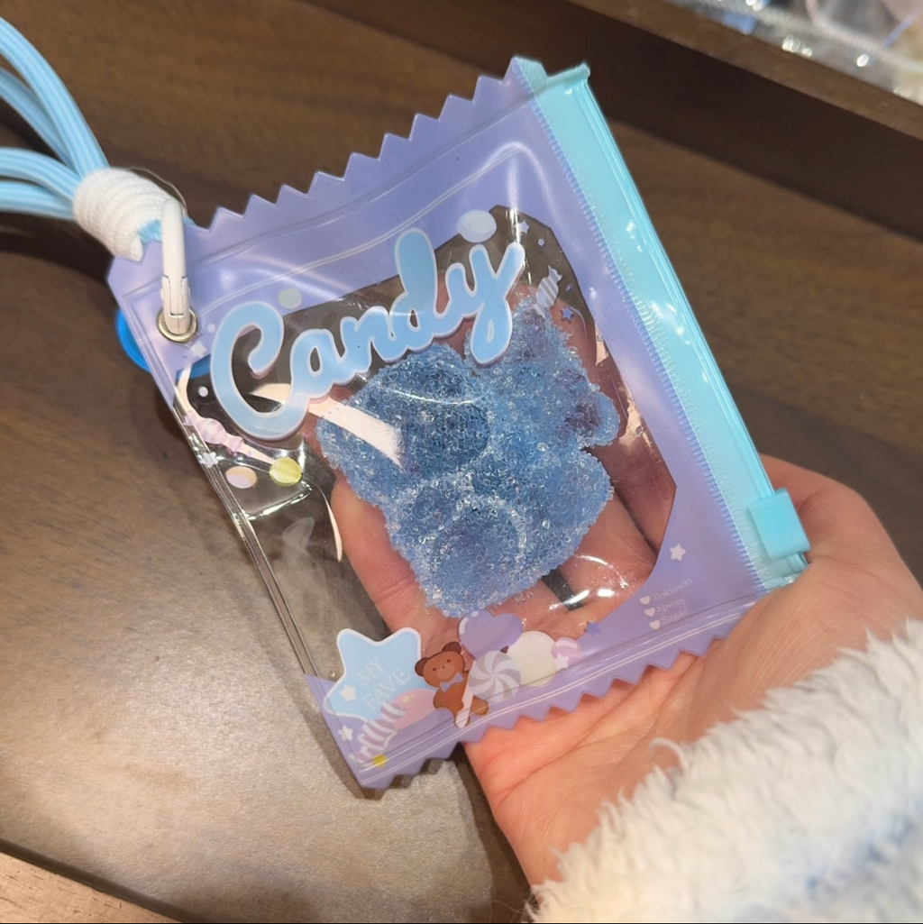 Jouet anti-stress ourson myrtille sable scintillant bleu tenu dans une main, sachet iridescent violet Candy zip bleu avec lanterne cordon bleu ciel, vue de face ambiance kawaii