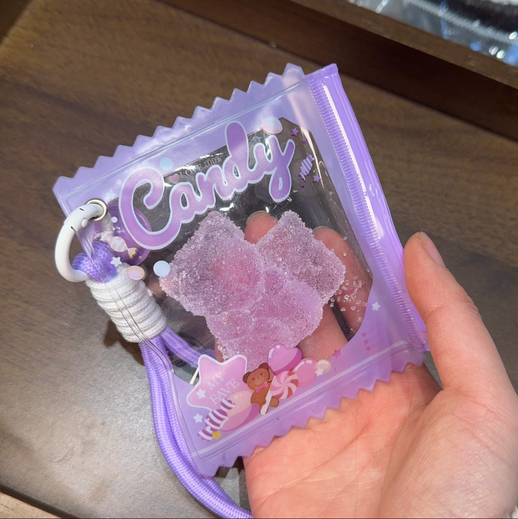 Jouet anti-stress ourson raisin sable scintillant rose-violet tenu dans une main, sachet violet lavé iridescent Candy zip violet avec cordon violet dégradé, posé sur table en bois, ambiance kawaii