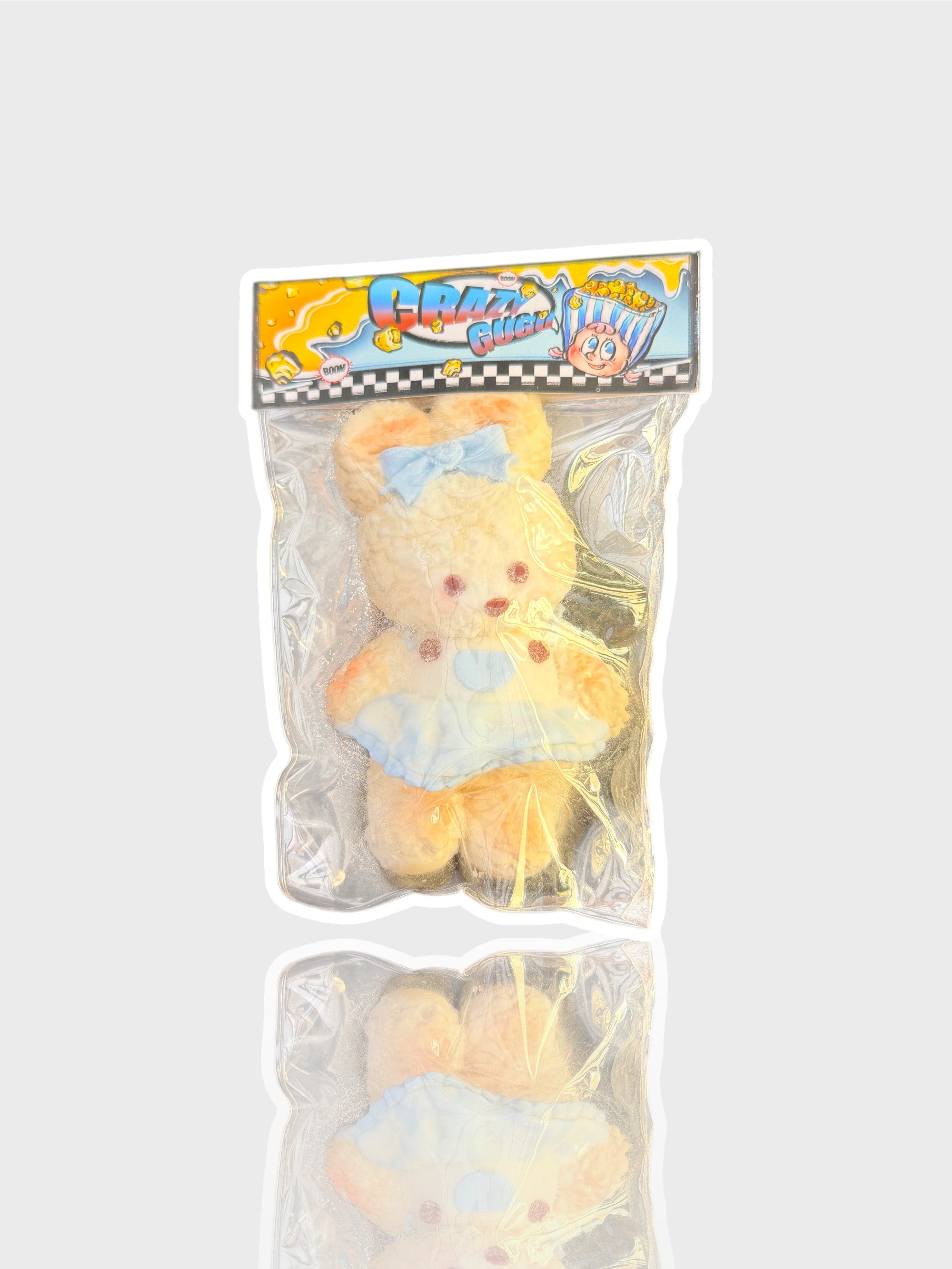 Jouet squishy peluche velours Crazy Gugu crème et bleu polyester ourson nœud papillon, vue de face dans emballage transparent fond blanc
