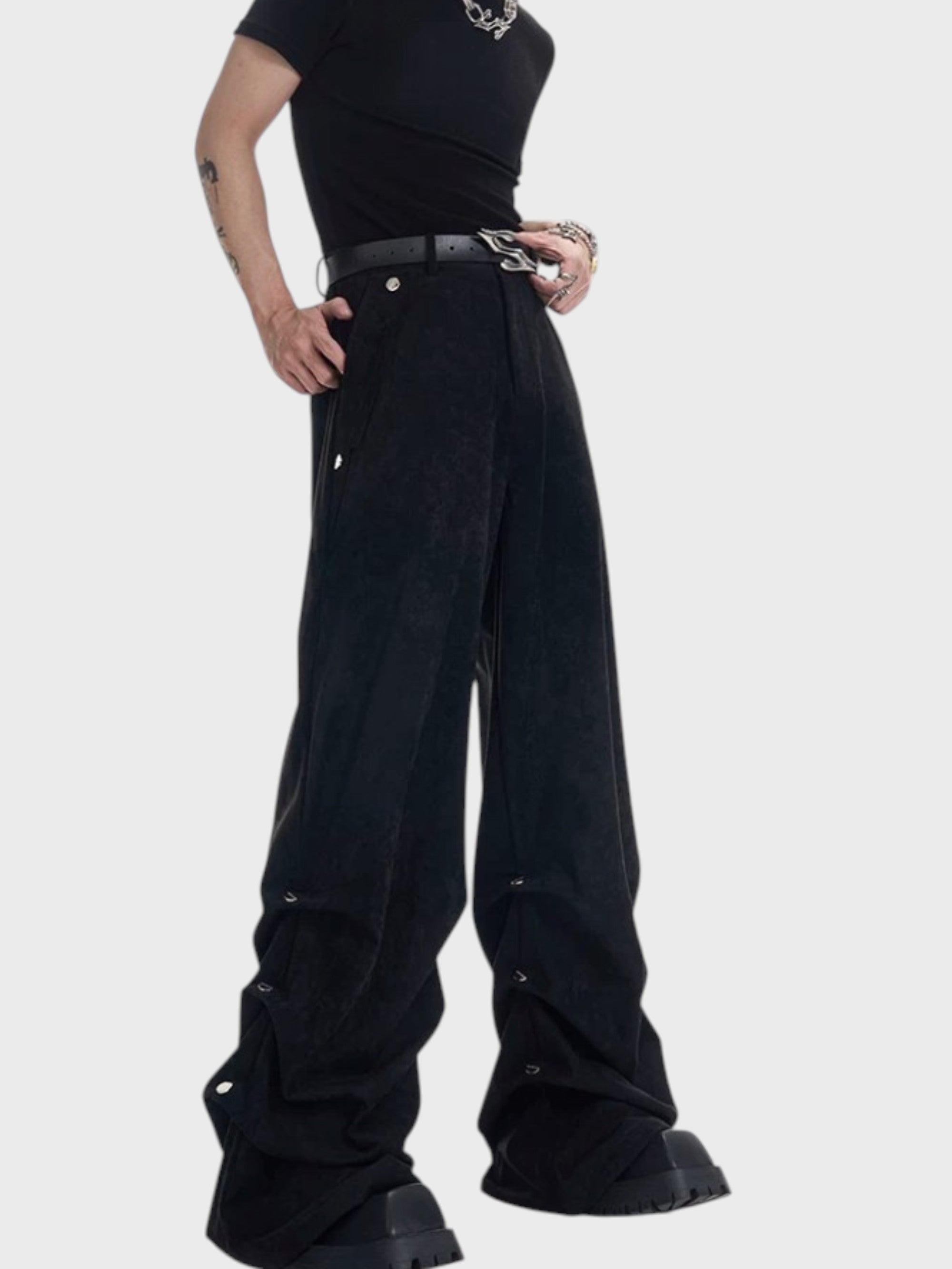 Pantalon large STARTEXCEEDEND velours suédé noir mat porté par un homme de profil, fronces accumulations bas jambes avec œillets argentés, ceinture boucle argentée et t-shirt noir, boots noires plateforme
