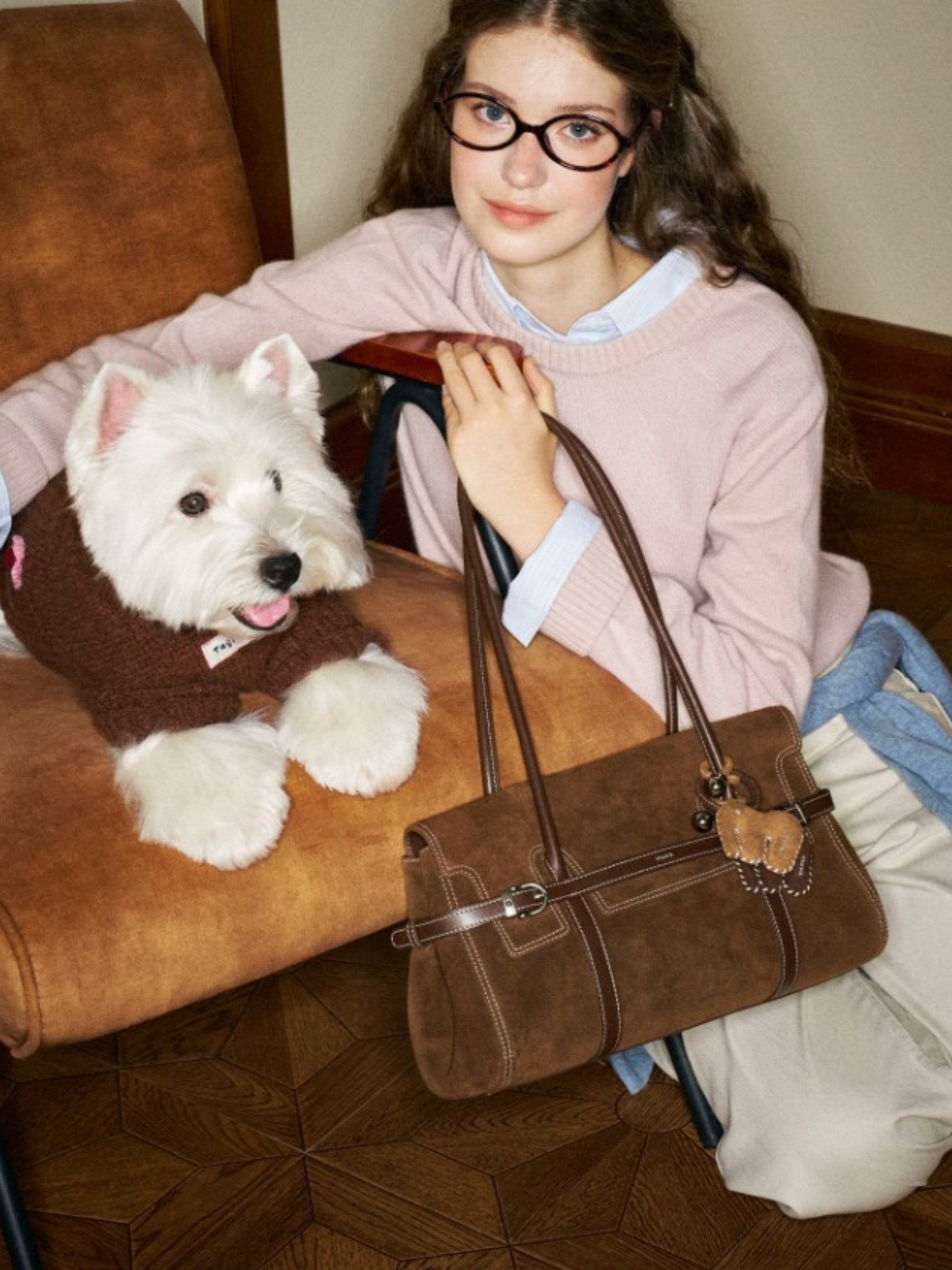 Sac baguette MUVA suédé marron chocolat porté à l'épaule par une femme aux lunettes ovales noires avec charm ours caramel, assise à côté d'un West Highland Terrier blanc, ambiance vintage preppy
