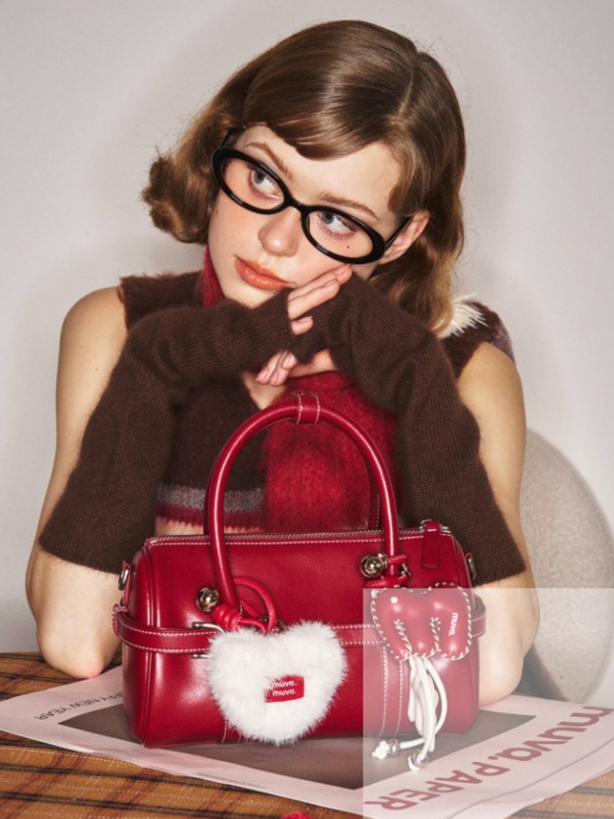 Sac boston MUVA cuir rouge bordeaux posé sur une table devant une femme aux lunettes ovales noires, charm cœur fourrure blanche avec logo MUVA attaché, ambiance vintage preppy
