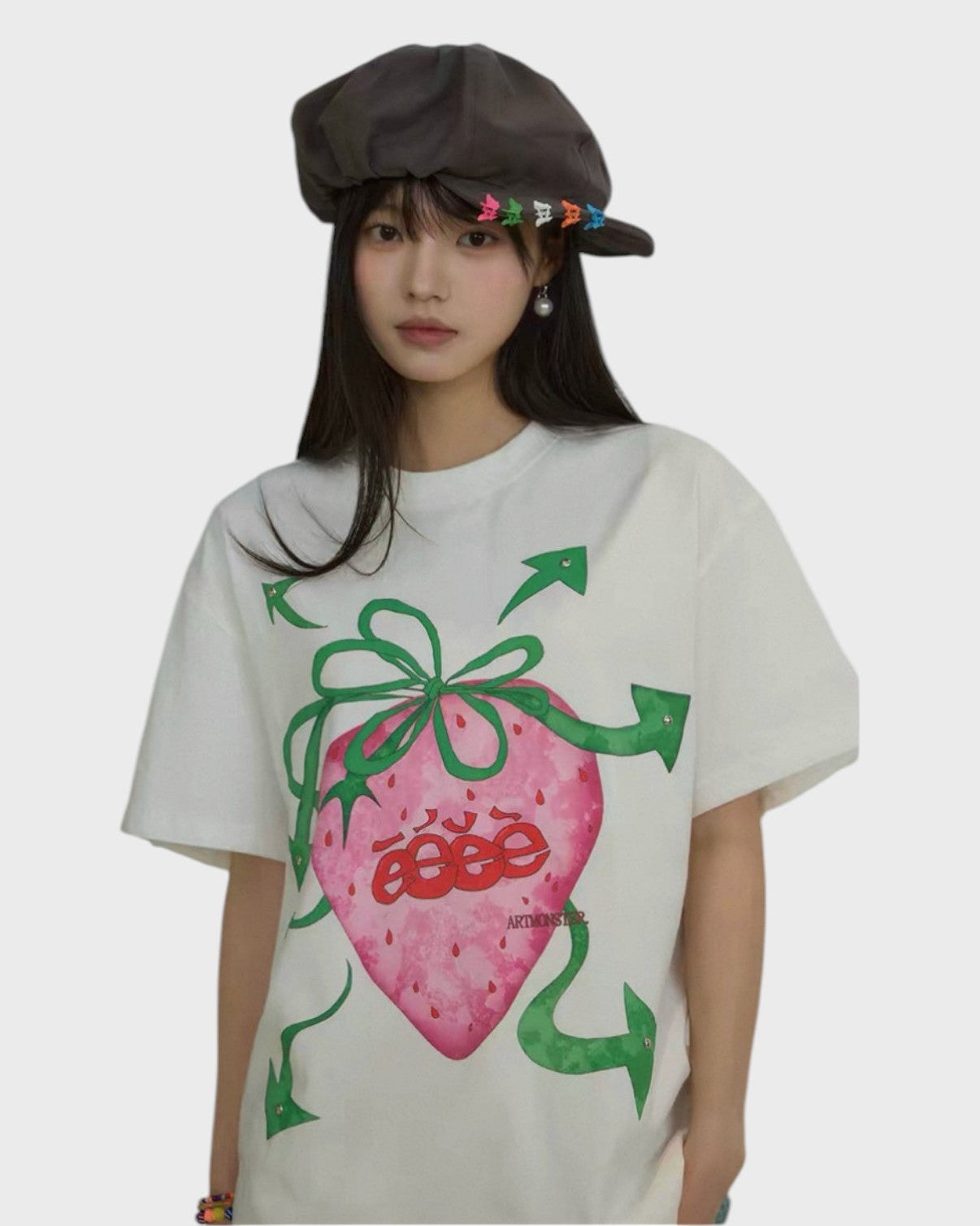 T-shirt oversize eeee Artmonster blanc imprimé fraise rose flèches vertes porté par une femme avec casquette baker boy marron et épingles colorées, vue de face