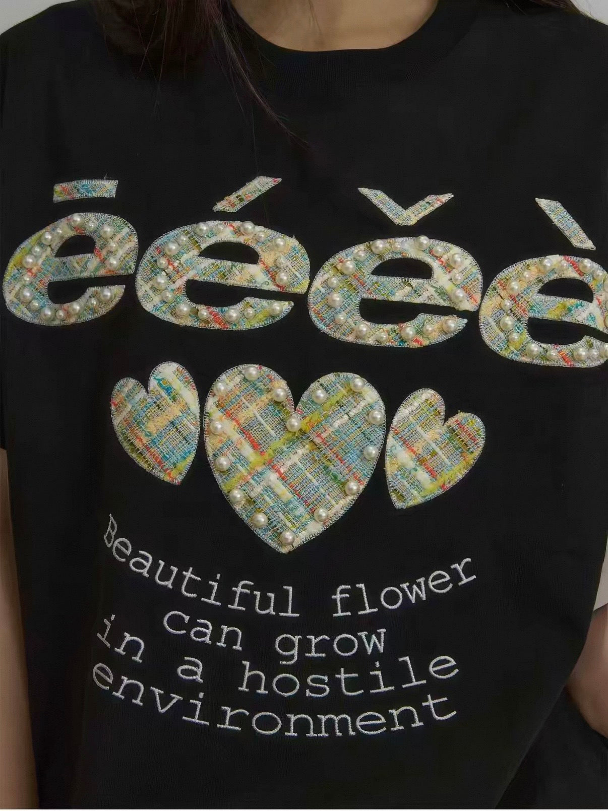 Gros plan sur les appliqués tweed multicolore et perles nacrées du t-shirt eeee Artmonster noir, logo eeee et trois cœurs brodés avec inscription "Beautiful flower can grow in a hostile environment"